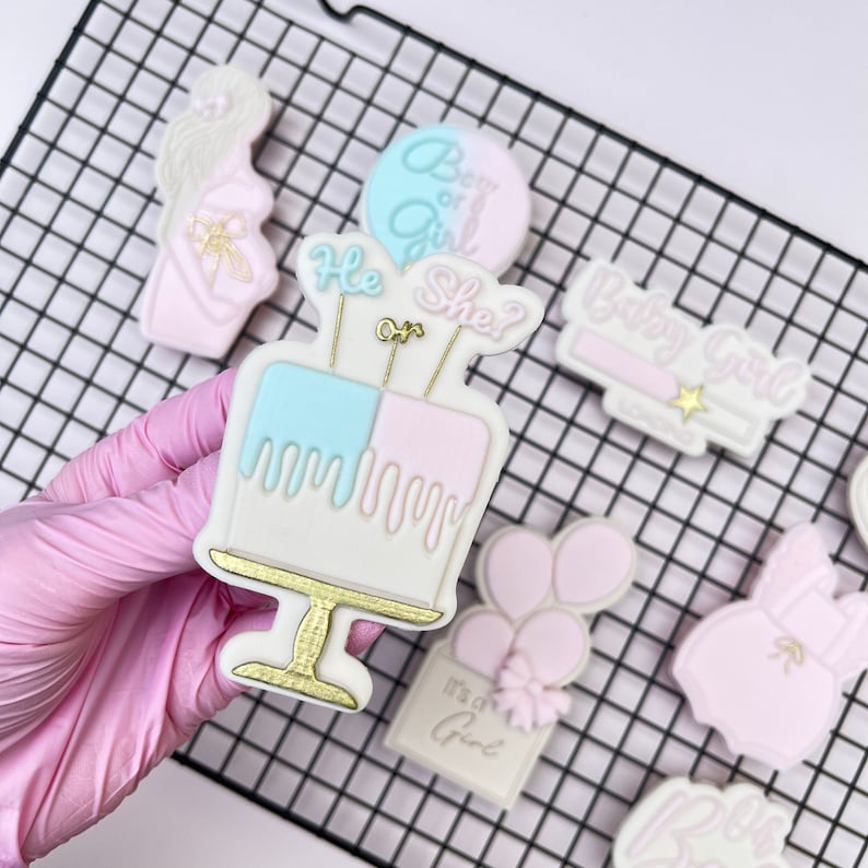 May include: A set of nine gender reveal cookies decorated with pink and blue icing. The cookies feature a cake, balloons, a baby outfit, and the phrases "Boy or Girl?", "It's a Girl", and "Baby Girl".