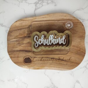 May include: A clear acrylic cookie cutter with a gold-toned border featuring the word Schulkind in a script font. The cutter is placed on a natural wood slab with the text CookiecutterFabrik and a circular logo printed on the wood surface.