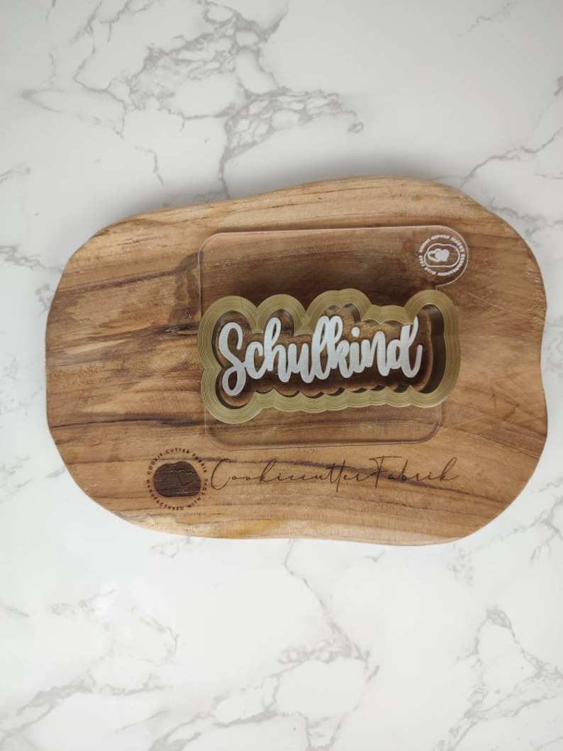May include: A clear acrylic cookie cutter with a gold-toned border featuring the word Schulkind in a script font. The cutter is placed on a natural wood slab with the text CookiecutterFabrik and a circular logo printed on the wood surface.