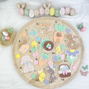 May include: A wooden tray displays Easter decorations. These include egg and bunny-shaped ornaments, a carrot patch sign, a nest with eggs, and a basket. The word "FROHEOSTERN" is spelled out with wooden letters.