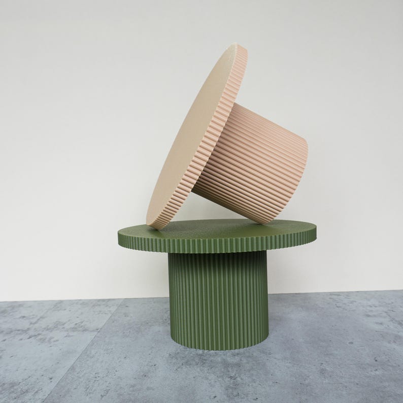 May include: A modern, abstract composition featuring three cylindrical tables. The bottom table is olive green, supporting a light pink table with a ridged edge. A third table, also light pink, rests on the second table. The tables have a minimalist design.