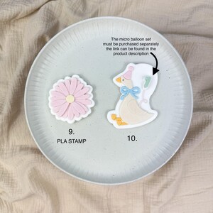 May include: Two cookie cutters on a plate. One is a pink daisy with a white border, and the other is a goose wearing a party hat and holding a balloon with the number 7. Text on the plate says "9. PLA STAMP" and "10."