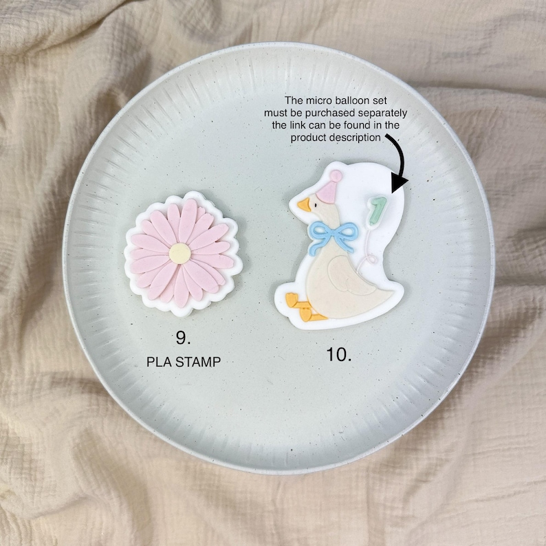 May include: Two cookie cutters on a plate. One is a pink daisy with a white border, and the other is a goose wearing a party hat and holding a balloon with the number 7. Text on the plate says "9. PLA STAMP" and "10."