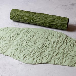 Könnte beinhalten: Ein grüner, strukturierter Nudelholz und eine passende Tonplatte. Das Nudelholz ist zylindrisch mit einem detaillierten, organischen Muster. Die Tonplatte zeigt das gleiche geprägte Design und bildet ein zusammenhängendes Set für Bastel- oder Backprojekte.
