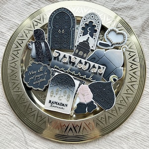 Könnte beinhalten: Eine Sammlung aufwendig dekorierter Kekse, angeordnet auf einer goldfarbenen Platte. Die Kekse weisen verschiedene Formen und Designs auf, darunter gewölbte Türen, Laternen und betende Hände. Einige Kekse tragen die Aufschriften "Ramadan Mubarak" und "Ramadan Kareem".