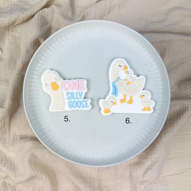 May include: Two whimsical goose-themed embellishments on a light blue plate. One says "ONE SILLY GOOSE" in pink and blue. The other shows a mother goose with two goslings. The plate sits on a beige fabric background.