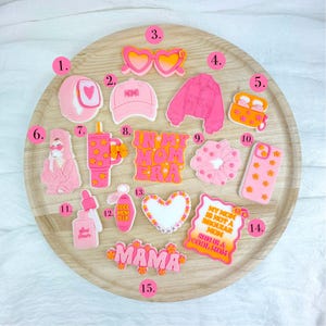 Op de afbeelding: Een verzameling roze en oranje accessoires, waaronder een zonnebril, een jasje, een telefoonhoesje en een scrunchie, gerangschikt op een houten dienblad. De items hebben bloemen- en hartontwerpen, met tekst als "In My Mom Era" en "Mama".