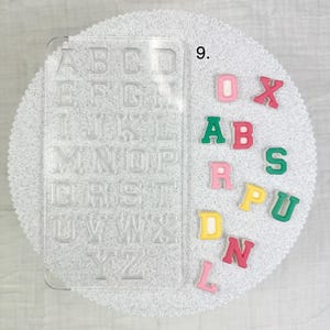 May include: A clear plastic alphabet mold with individual letter spaces, next to colorful foam letters. The letters are in various colors, including pink, green, yellow, and red. The mold is on a white, textured surface.