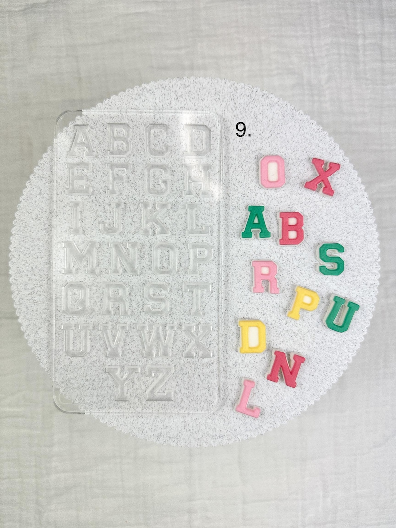 May include: A clear plastic alphabet mold with individual letter spaces, next to colorful foam letters. The letters are in various colors, including pink, green, yellow, and red. The mold is on a white, textured surface.