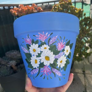 Peut inclure: Un pot de fleurs en terre cuite bleu avec un motif peint à la main de marguerites blanches et de fleurs violettes.