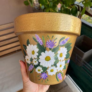 Peut inclure: Un pot de fleurs en terre cuite doré avec un motif peint à la main de marguerites blanches et de lavande violette.
