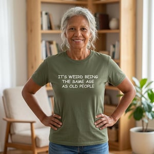 May include: An olive green short-sleeved crew neck t-shirt featuring the white text "IT'S WEIRD BEING THE SAME AGE AS OLD PEOPLE" printed across the chest. The garment is made of a solid-colored fabric and has a standard fit with a straight hemline.