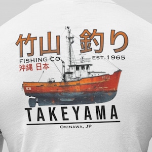 Op de afbeelding: Wit langarm shirt met een rode en witte visboot afbeelding. De boot heeft de tekst "竹山 釣り" en "FISHING CO. 沖繩日本" aan de zijkant. De tekst "TAKEYAMA" en "OKINAWA, JP" staat onder de boot.