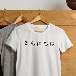 Puede incluir: Tres camisetas colgadas de perchas sobre un fondo de madera. La camiseta de la derecha es blanca con texto japonés negro que dice "こんにち は".