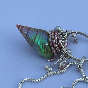 Puede incluir: Un collar artesanal con un colgante de concha iridiscente. El colgante está adornado con perlas rosas y grises, y detalles de alambre de plata. Una delicada cadena de plata completa el diseño, creando una joya única y elegante.