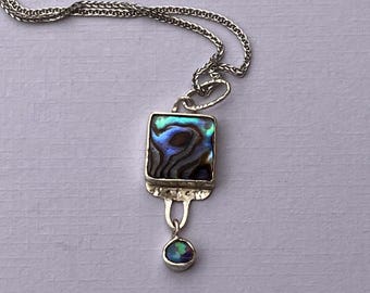 May include: A silver necklace featuring a square pendant with an iridescent abalone shell inlay. Below the square pendant hangs a small, round, iridescent stone. The necklace chain is a delicate, double strand design.