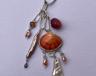 May include: A silver necklace with a pendant incorporating an orange seashell, pearls, and a mother-of-pearl hand charm. Other charms include a red spiral bead and a small green bead. The necklace is suspended from a fine chain.