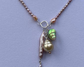 May include: A necklace with a strand of small, light brown beads. The pendant features a long, pinkish-beige pearl, a cream-coloured pearl, and a green, iridescent shell-shaped bead. This unique necklace is a piece of jewellery.