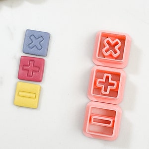 May include: Three sets of plastic cookie cutters in the shapes of a plus sign, minus sign, and multiplication sign. The cutters are in pink, red, yellow, and blue.