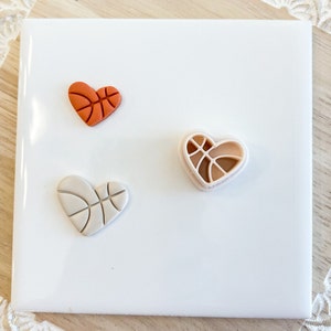May include: Three heart-shaped clay ornaments with a basketball design. One is orange, one is white, and one is a light peach color.