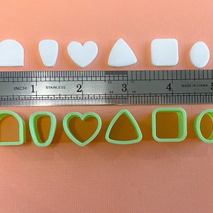 May include: Set of eight white and green plastic cookie cutters in various shapes, including an arch, oval, heart, triangle, square, and circle. The cutters are arranged in two rows on a pink surface with a ruler for scale.