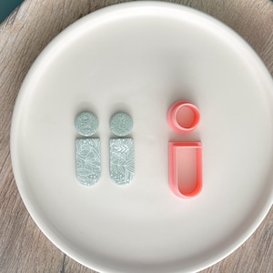 May include: Two pairs of light blue polymer clay earrings with a white floral pattern. The earrings are displayed on a white plate with two pink polymer clay cutters.