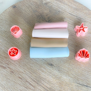 May include: Four silicone placemats in blue, beige, white, and pink are stacked on a wooden surface. Four pink plastic cookie cutters shaped like seashells and starfish are scattered around the placemats.