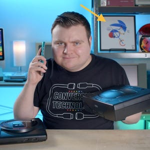 May include: A black Sega Genesis video game console sits on a wooden surface, while a person holds a second black console. A framed Sonic the Hedgehog illustration is visible on a shelf behind, along with a computer monitor displaying a game.