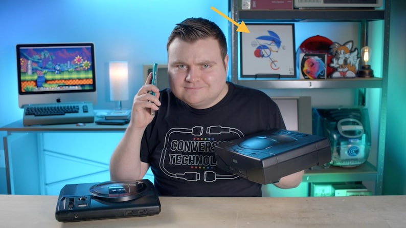 May include: A black Sega Genesis video game console sits on a wooden surface, while a person holds a second black console. A framed Sonic the Hedgehog illustration is visible on a shelf behind, along with a computer monitor displaying a game.