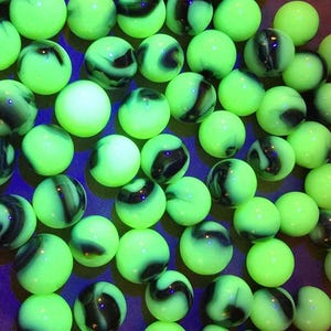 May include: A collection of green marbles with black and white swirls. The marbles have a glowing appearance, and are scattered across a surface. The marbles are a classic toy, and are often used for games and collecting.