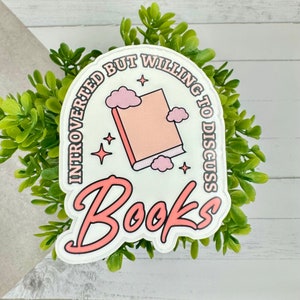 Könnte beinhalten: Ein weißer Aufkleber mit rosa Text und einer rosa Buchillustration. Der Text lautet "Introvertiert, aber bereit, über Bücher zu diskutieren".