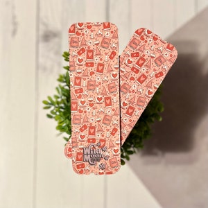 May include: Two pink bookmarks with a repeating pattern of hearts, stars, books, and potion bottles. The bookmark on the left has the text "Wild Moon Magic" in a pink, purple, and white font.