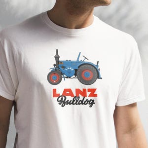 Puede incluir: Camiseta blanca con un diseño de tractor azul y detalles rojos. El diseño del tractor incluye la palabra "LANZ" en un rectángulo rojo encima de la palabra "Bulldog" en una fuente estilizada. El diseño del tractor está centrado en la camiseta.