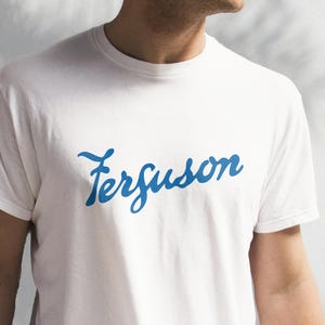 May include: White t-shirt with a blue cursive script that says "Ferguson" on the front.