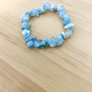 Pode incluir: Uma pulseira de lascas de água-marinha azul clara. A pulseira é feita de pequenas pedras de água-marinha de formato irregular, amarradas em uma faixa elástica. As pedras variam em tons de azul e branco. A pulseira é exibida em uma superfície de madeira marrom clara.