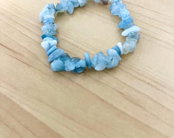 Puede incluir: Un brazalete de virutas de aguamarina azul claro. El brazalete está hecho de pequeñas piedras de aguamarina de forma irregular ensartadas en una banda elástica. Las piedras varían en tonos de azul y blanco. El brazalete se exhibe sobre una superficie de madera marrón claro.