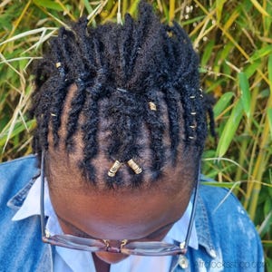 May include: Close-up of black dreadlocks adorned with gold-colored hair beads. The hair is styled in a neat pattern, with the beads adding a decorative touch. The image is taken from a high angle, with a background of green foliage.