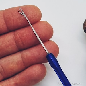 Op de afbeelding: Een close-up van een zilveren metalen latch hook tool met een blauwe handgreep. De haak heeft een gebogen uiteinde met een gespleten punt. De tool is ontworpen voor haarvlechten en wordt in een hand gehouden.