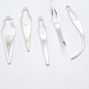 Puede incluir: Cinco agujas de crochet plateadas, cuatro son puntiagudas y una es curva. Las agujas están sobre una superficie blanca.