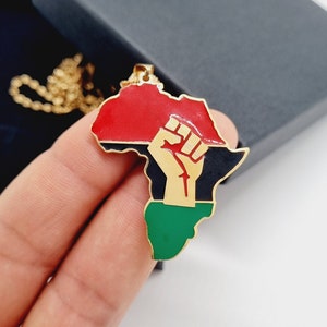 K&ouml;nnte beinhalten: Goldkette mit einem Anh&auml;nger in Form des afrikanischen Kontinents. Der Anh&auml;nger ist in den Farben der panafrikanischen Flagge bemalt: rot, schwarz und gr&uuml;n. Eine erhobene goldene Faust befindet sich in der Mitte des Anh&auml;ngers.