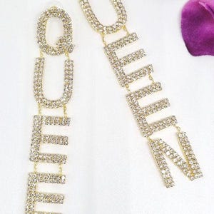 May include: A pair of gold-tone earrings with the word "QUEEN" spelled out in rhinestones. The earrings are long and dangle.