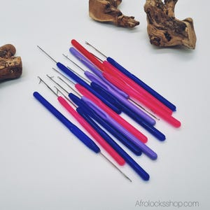 May include: A collection of colorful crochet hooks in shades of blue, pink, purple, and red. The hooks have metal tips and plastic handles. The image is taken from a top-down perspective, with the hooks arranged on a white surface.