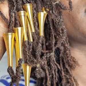 May include: Gold metal hair beads in a conical shape, attached to brown dreadlocks. The beads are about 1 inch long.