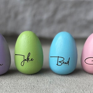 May include: Seven wooden Easter eggs painted in pastel colors with names written on them: Dad, Vivian, Jake, Brad, Carly, and Mia.