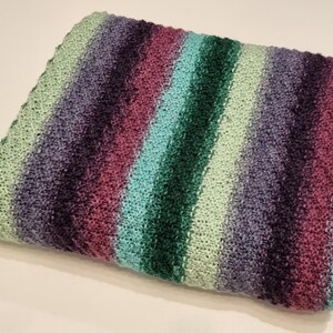 May include: Folded knitted blanket with a striped pattern. The blanket features bands of colour including light green, purple, teal, dark green, and magenta. The blanket is folded neatly and appears soft.