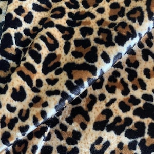 Op de afbeelding: Close-up van een stof met een luipaardprint. De stof heeft een beige achtergrond met zwarte en bruine vlekken. De stof is gevouwen, waardoor de textuur en het patroon zichtbaar zijn.