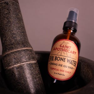 May include: A brown glass bottle with a spray nozzle labeled "Evil Bone Water" sits in a dark gray mortar. The label reads "Saint Apothecary" and "Topical Use Only". The mortar and pestle are made of dark gray stone.