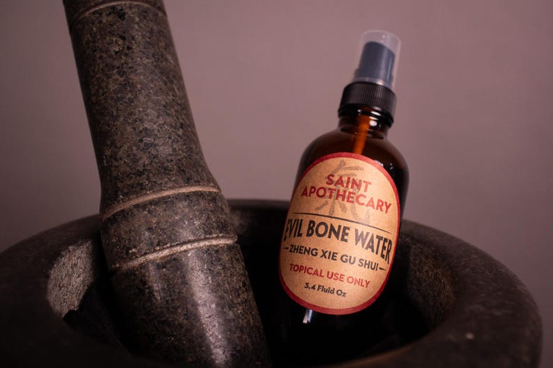 May include: A brown glass bottle with a spray nozzle labeled "Evil Bone Water" sits in a dark gray mortar. The label reads "Saint Apothecary" and "Topical Use Only". The mortar and pestle are made of dark gray stone.