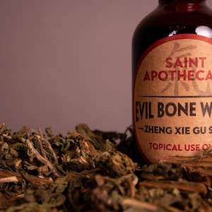 May include: A brown glass bottle with a red and tan label that reads "Saint Apothecary Evil Bone Water." The bottle is surrounded by dried herbs and plant matter. The text "Topical Use Only" is visible on the label.