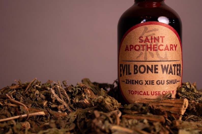 May include: A brown glass bottle with a red and tan label that reads "Saint Apothecary Evil Bone Water." The bottle is surrounded by dried herbs and plant matter. The text "Topical Use Only" is visible on the label.
